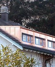 Weißes Haus mit Schornsteinen, grüne Fensterläden, Wald im Hintergrund.