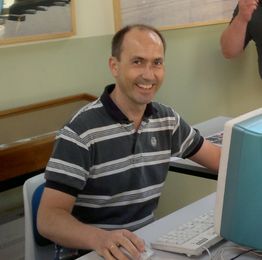 Ein Mann sitzt lächelnd vor einem Computer in einem Raum mit hellen Wänden.