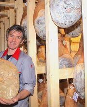 Mann hält großen Schinken in einem Lagerraum voller hängender Schinkenstücke.