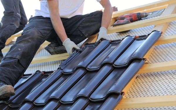 Ein Dachdecker sitzt auf dem Dachfirst und Deckt das Dach mit anthrazitfarbenen Dachziegeln.