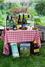 Flaschen und Boxen mit Wein und Öl auf kariertem Tuch im Garten, umgeben von Paprika und Zitrone.