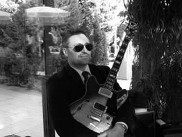 Mann in Anzug mit Sonnenbrille hält eine E-Gitarre. Er sitzt im Freien vor einer Glaswand.