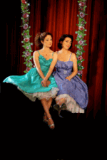 Zwei Frauen in bunten Kleidern auf einer Schaukel vor rotem Vorhang, umrahmt von Blumen.