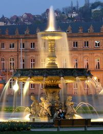 Brunnen im Schlossplatz mit beleuchtetem Gebäude im Hintergrund, zwei Menschen auf einer Bank.