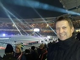 Ein Mann lächelt in einem Stadion bei Nacht mit Lichtern und Zuschauern im Hintergrund.