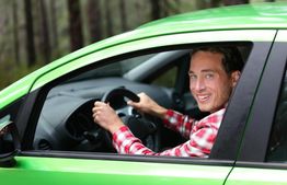 Man in a green car, wearing a red plaid shirt, smiling through the driver’s side window.