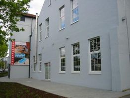 Graues Gebäude mit Reklame für Eröffnung an der Seite, daneben ein Baum und Gehweg.