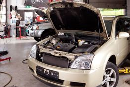 Car in a garage with an open hood, another vehicle in the background being serviced.