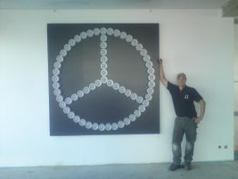 Mann steht neben großem Mercedes-Logo an Wand, aus kleinen Autologos geformt.
