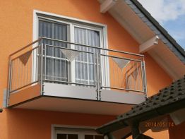 Balkon mit Metallgeländer an orangefarbenem Haus, darüber Fenster mit weißen Vorhängen.