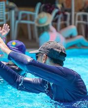 Pessoas praticando hidroginástica em piscina azul, com cadeiras e chapéus ao fundo.
