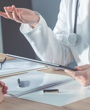 Ein Arzt mit Stethoskop spricht mit einem Patienten. Auf dem Tisch liegen Unterlagen und eine Brille.