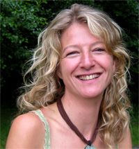 Eine lächelnde Frau mit lockigem Haar im Freien vor einem Hintergrund von grünen Bäumen.