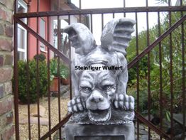 Steinfigur eines geflügelten Dämons vor einem Zaun, umgeben von Pflanzen und einem Haus im Hintergrund.