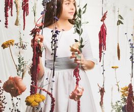 Niña con corona de flores, rodeada de flores suspendidas, mira hacia la derecha.