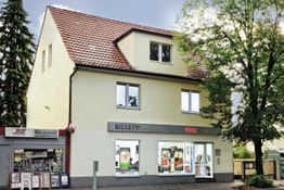 Zweistöckiges Gebäude mit Geschäften im Erdgeschoss und Bäumen an der Seite.