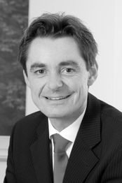 Schwarz-weiß Porträt eines lächelnden Mannes in Anzug und Krawatte vor einem unscharfen Hintergrund.