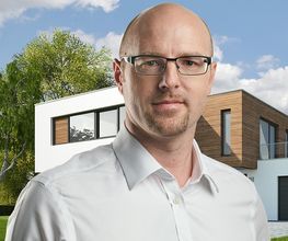 Ein Mann mit Brille vor einem modernen Haus, Bäume im Hintergrund, sonniger Himmel.