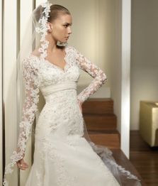 Sposa in abito bianco con velo e maniche in pizzo, appoggiata a una parete interna.