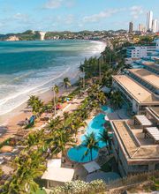 Vista aérea de uma praia com edifícios ao lado e morros ao fundo em um dia ensolarado.