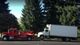 A red tow truck pulls a white box truck on a road with tall evergreen trees in the background.