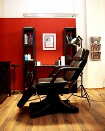 Barbershop-Stuhl vor roter Wand, flankiert von Regalen und Lesematerialien, Lampenbeleuchtung.
