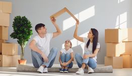 Family sitting with a cardboard roof shape over their heads, surrounded by moving boxes.