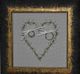 Stickbild mit Herzmotiv, zwei Ringen und Schleifen, in einem antiken Rahmen.