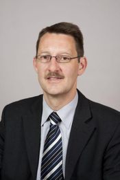 Ein Mann mit Brille in einem Anzug und gestreifter Krawatte vor grauem Hintergrund.