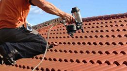 Worker using a nail gun on a red tiled roof under a blue sky.