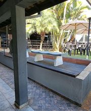 Corredor com mesas e cadeiras ao ar livre, cobertura de lona e palmeiras ao fundo.