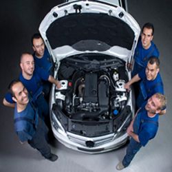 Six mechanics standing around a car with an open hood.