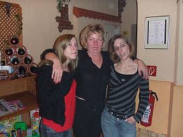 Drei Frauen posieren eng umarmt in einem gemütlichen Raum mit Holzelementen und Weinregalen.