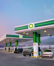 Estación de gasolina con varios surtidores y coches al atardecer, montaña al fondo.