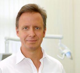 Eine Person in weißem Hemd lächelt in einem Raum mit zahnärztlicher Beleuchtung im Hintergrund.