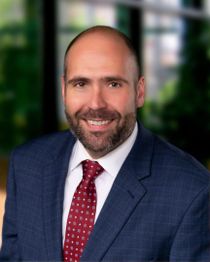Smiling man in a suit with a red tie stands in front of a blurred indoor background.