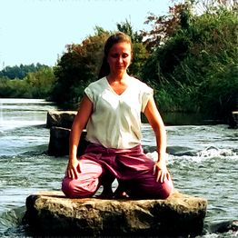 Eine Frau meditiert in rosa Hose auf einem Felsen im Fluss, umgeben von Natur.