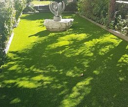 Garten mit grünem Rasen, Sonnenschein, Palme und einer kleinen Statue in der Mitte.