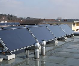 Solarmodule auf einem Flachdach in einer Wohngegend, mit Häusern und Bäumen im Hintergrund.