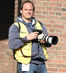 Ein Mann mit gelber Weste hält eine Kamera und steht vor einer Ziegelwand im Freien.