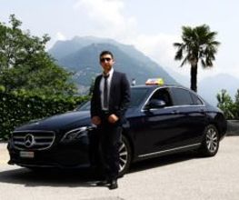 Ein Mann in Anzug steht vor einem schwarzen Mercedes auf einer Straße mit Berg- und Palmenhintergrund.