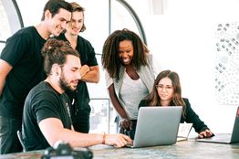 Group of five people smiling and collaborating around a laptop in a bright office space.