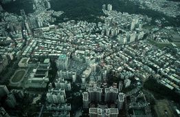 Luftaufnahme einer dichten urbanen Landschaft mit vielen Gebäuden und einem grünen Hügel im Hintergrund.