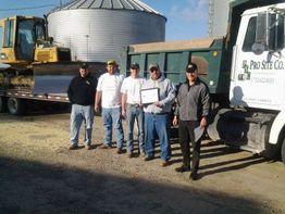 Five men standing beside a truck and bulldozer, with one holding a certificate, on a sunny day.
