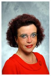 Frau mit Brille und rotem Oberteil vor grauem Hintergrund, schaut freundlich in die Kamera.