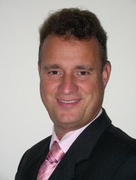 Ein Mann in Anzug und Krawatte lächelt in die Kamera, mit hellem Hintergrund.