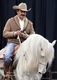 Man in cowboy attire riding a white horse indoors.