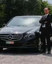 Mann in Anzug steht mit verschränkten Armen vor einem schwarzen Mercedes auf Parkplatz.