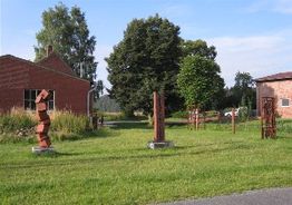 Skulpturen aus Metall stehen auf einer Wiese, umgeben von Bäumen und roten Backsteingebäuden.