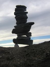 Ein Stapel flacher Steine balanciert auf sandigem Boden, vor einem wolkigen Himmel.